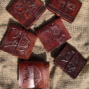 May include: A collection of six brown leather journals with embossed designs. Each journal features a unique image, including trees and elephants, and is secured with a matching leather tie. The journals are arranged on a textured, tan fabric surface.