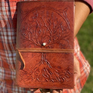 May include: A brown leather journal with a tree of life design embossed on the cover. The tree has intricate branches and roots, and a small white circle is in the center of the tree. The journal has a brown leather strap closure.