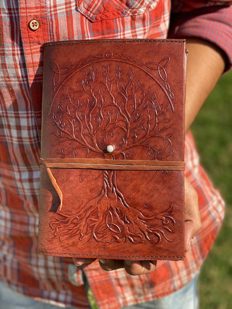 May include: A brown leather journal with a tree of life design embossed on the cover. The tree has intricate branches and roots, and a small white circle is in the center of the tree. The journal has a brown leather strap closure.