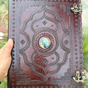 May include: A handcrafted, burgundy leather journal with intricate embossed patterns and a central gemstone. The journal features a decorative metal clasp and stitched edges. The cover design includes floral and geometric elements, creating a vintage aesthetic.