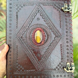 May include: A dark brown leather journal with intricate embossed patterns and a central diamond design. The journal features a gemstone and decorative metal clasps. The edges are stitched, and the overall aesthetic is rustic and handcrafted.