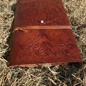 May include: A brown leather journal with a tooled design of a tree of life with a Celtic knot at the base. The journal has a leather strap closure and a metal button.