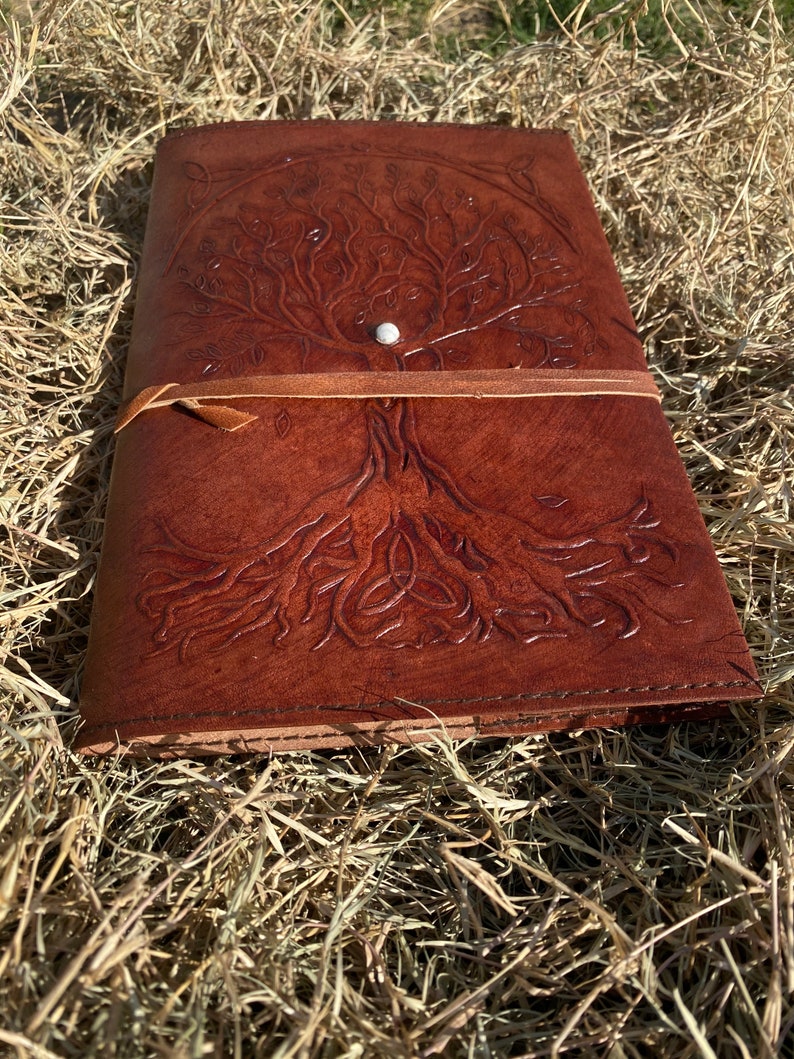 May include: A brown leather journal with a tooled design of a tree of life with a Celtic knot at the base. The journal has a leather strap closure and a metal button.