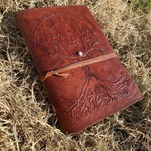 May include: A brown leather journal with a tree of life design embossed on the cover. The journal has a leather strap closure and a silver button.