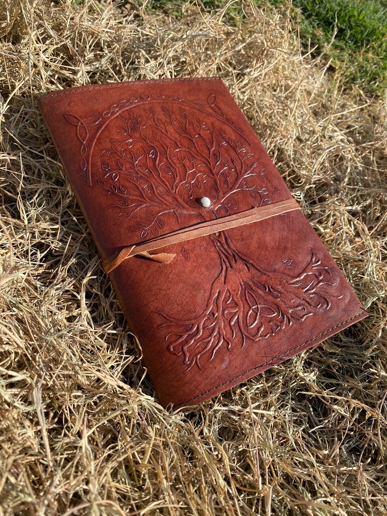 May include: A brown leather journal with a tree of life design embossed on the cover. The journal has a leather strap closure and a silver button.