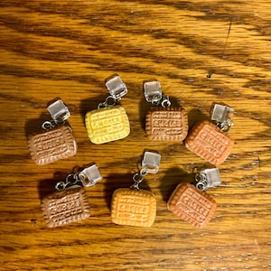 Puede incluir: Una colección de dijes en miniatura con forma de galleta en varios tonos de marrón y amarillo. Cada dije tiene la palabra "CHOICE" impresa. Los dijes están unidos a pequeños aros plateados con cierres de plástico transparente.