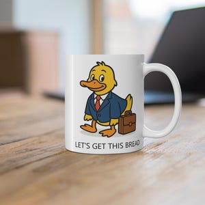 Könnte beinhalten: Weiße Keramik-Tasse mit einer Cartoon-Ente in einem blauen Anzug, roter Krawatte und Aktentasche. Auf der Tasse steht der Text "LET'S GET THIS BREAD". Die Tasse steht auf einer Holzoberfläche.