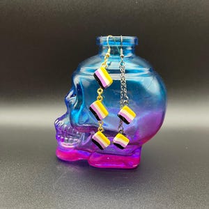 May include: Non-binary pride flag earrings. The earrings feature small, square charms in the colors of the non-binary flag: yellow, white, purple, and black. One earring has gold-colored hooks and chain links, while the other has silver-colored chain links.