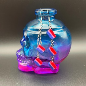 May include: Silver chain earrings with rectangular charms. Each charm is a combination of red, white, and blue. The earrings are displayed on a blue skull-shaped glass bottle with a pink and purple base.