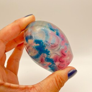 May include: An oval-shaped decorative object with a colorful, abstract design. The object features swirls of blue, pink, and silver glitter against a clear background. The object is held in a hand against a white background.