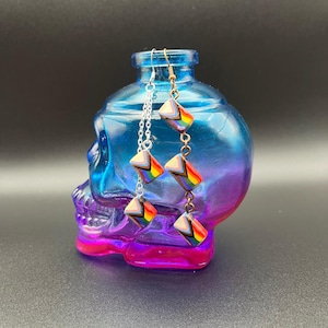 May include: Earrings with a rainbow geometric design hang in front of a blue and purple skull-shaped glass container. The earrings have a silver chain and gold-colored hooks. The earrings are made of a colorful material.