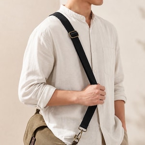 May include: A black adjustable crossbody bag strap featuring a metal slider and a swivel lobster clasp hook, attached to an olive green canvas bag, worn over a light beige long-sleeved button-down shirt with a mandarin collar.