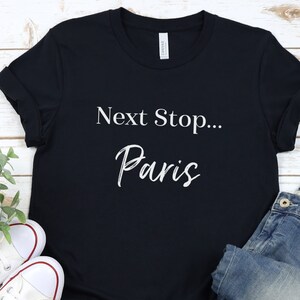 May include: A black t-shirt with white text that reads "Next Stop... Paris". The shirt is laid flat on a white wooden surface with a pair of white trainers and blue jeans.