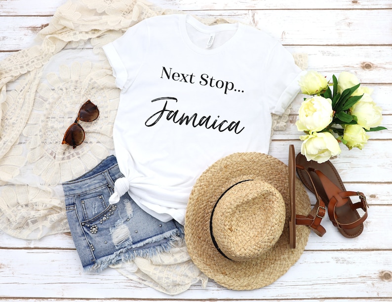 May include: A white t-shirt with the text 'Next Stop... Jamaica' in black lettering. The shirt is laid out on a white wooden surface with a pair of denim shorts, a straw hat, a pair of brown sandals, and a white lace shawl.