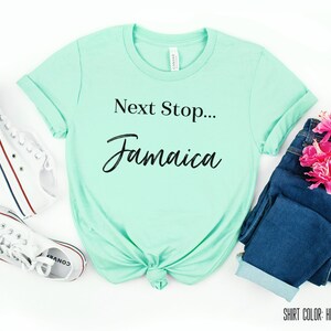 May include: A heather mint t-shirt with the text "Next Stop... Jamaica" in black lettering. The shirt is laid flat on a white surface with a pair of white trainers, blue jeans, and pink flowers.