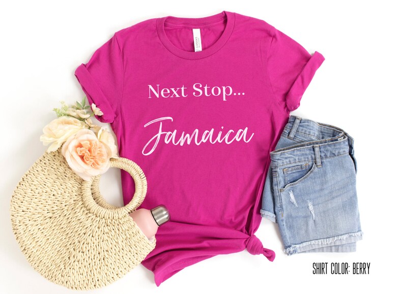 May include: A pink t-shirt with the text "Next Stop... Jamaica" in white. The shirt is laid flat on a white background with a straw bag, a water bottle, and denim shorts.