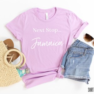 May include: Lilac t-shirt with the text "Next Stop... Jamaica" in white. The shirt is styled with denim shorts, a straw bag, sandals, and sunglasses.