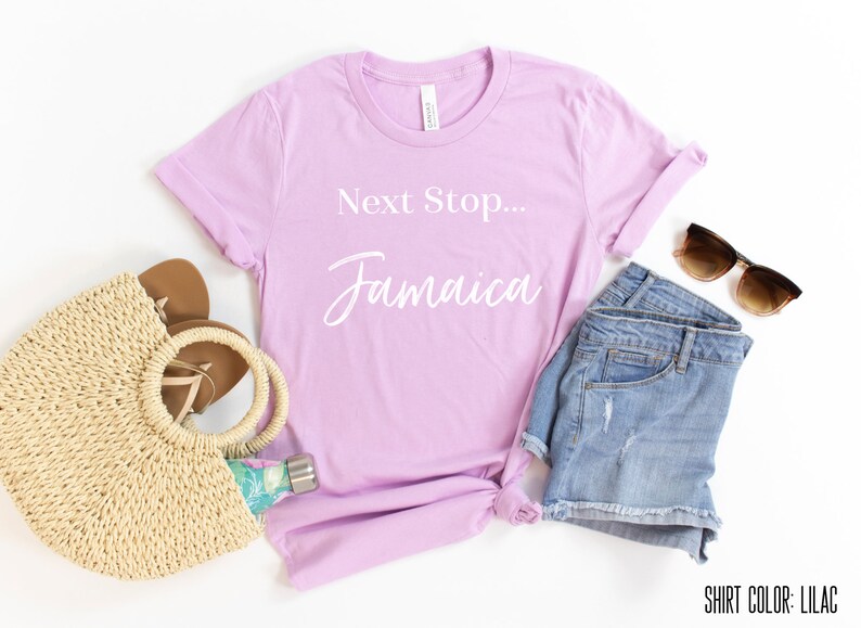May include: Lilac t-shirt with the text "Next Stop... Jamaica" in white. The shirt is styled with denim shorts, a straw bag, sandals, and sunglasses.
