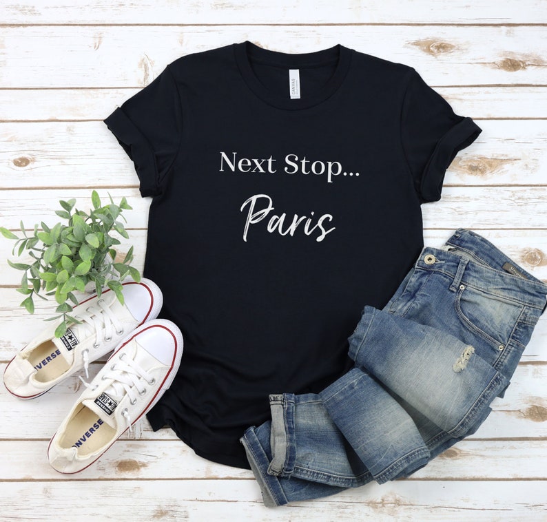May include: A black t-shirt with white text that reads "Next Stop... Paris". The shirt is laid flat on a white wooden surface with a pair of white trainers and blue jeans.