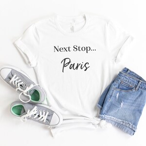 May include: White t-shirt with the text "Next Stop... Paris" in black lettering. The shirt is laid flat on a white surface with a pair of grey sneakers and denim shorts.