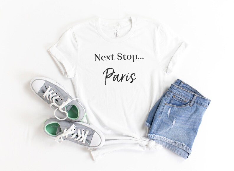 May include: White t-shirt with the text "Next Stop... Paris" in black lettering. The shirt is laid flat on a white surface with a pair of grey sneakers and denim shorts.