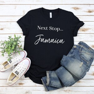 May include: Black t-shirt with white text that reads "Next Stop... Jamaica". The shirt is laid flat on a white wood background with a pair of white trainers and blue jeans.
