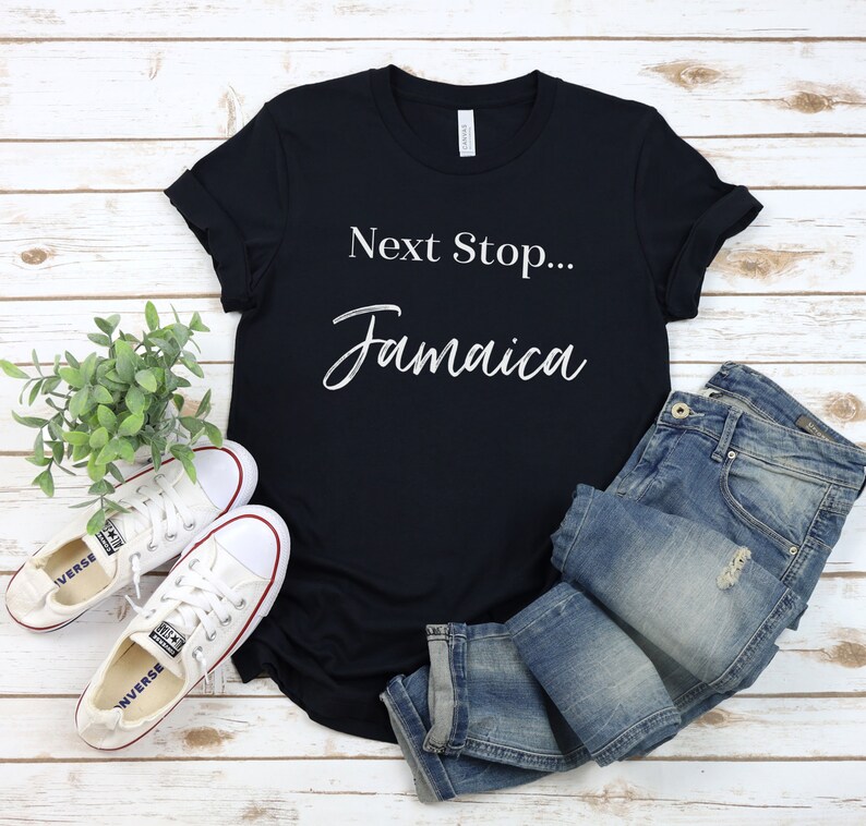 May include: Black t-shirt with white text that reads "Next Stop... Jamaica". The shirt is laid flat on a white wood background with a pair of white trainers and blue jeans.