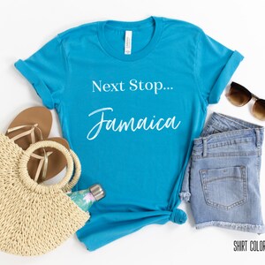 May include: Aqua blue t-shirt with the text "Next Stop... Jamaica" in white. The shirt is laid flat on a white surface with a straw bag, sandals, denim shorts, and sunglasses.