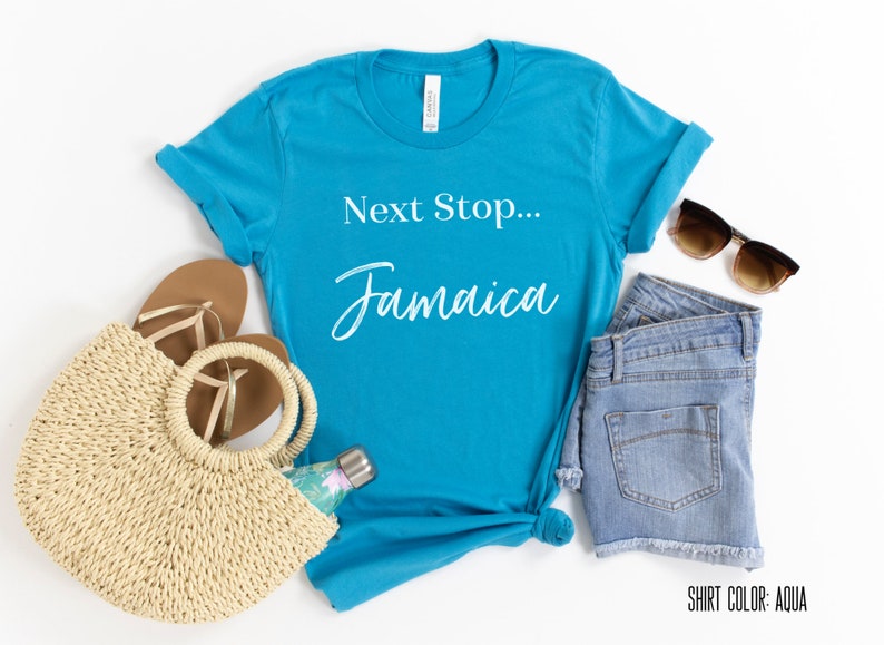 May include: Aqua blue t-shirt with the text "Next Stop... Jamaica" in white. The shirt is laid flat on a white surface with a straw bag, sandals, denim shorts, and sunglasses.