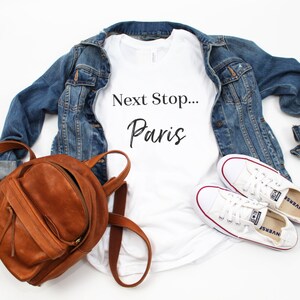 May include: White t-shirt with the text "Next Stop... Paris" in black lettering. The shirt is paired with a denim jacket, a brown leather backpack, and white trainers.