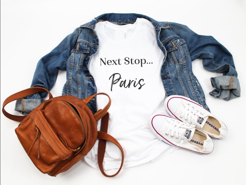 May include: White t-shirt with the text "Next Stop... Paris" in black lettering. The shirt is paired with a denim jacket, a brown leather backpack, and white trainers.