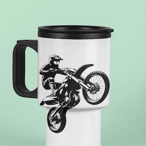 May include: A white travel mug with a black lid and handle. The mug features a black and white silhouette of a motocross rider performing a stunt.