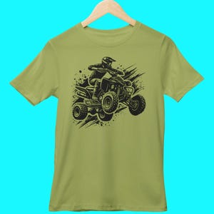 May include: A green t-shirt with a black graphic of a person riding an ATV. The ATV is surrounded by black splatters of paint.
