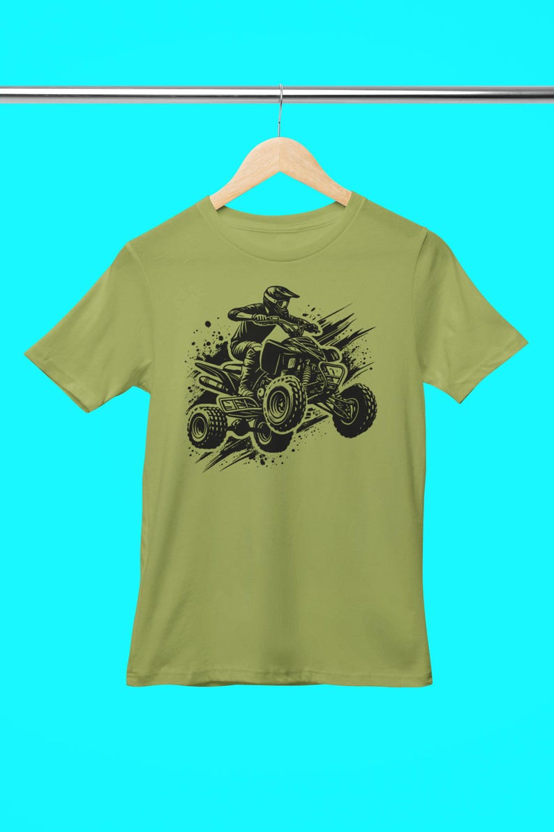 May include: A green t-shirt with a black graphic of a person riding an ATV. The ATV is surrounded by black splatters of paint.