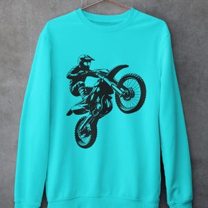 May include: A turquoise blue sweatshirt with a black silhouette of a motocross rider performing a wheelie on their dirt bike.