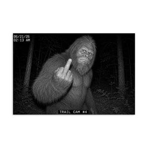 May include: A black and white trail camera photograph of a hairy, ape-like creature in a forest. The creature is holding up its middle finger. Text in the corner reads 06/21/26 02:13 AM and TRAIL CAM #4 at the bottom of the frame.