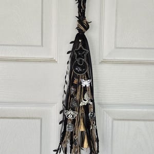 May include: A decorative black and tan tassel with various charms, including stars, butterflies, and bells. The tassel is adorned with black ribbon and hanging from a twisted cord with faux leaves. A unique, handcrafted item.