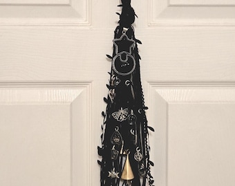 May include: A black fabric tassel wall hanging featuring various silver-toned metal charms including a star, a moth, a planchette, and a pentagram, along with a single gold-toned bell charm, all suspended from a black fabric loop with leaf accents.