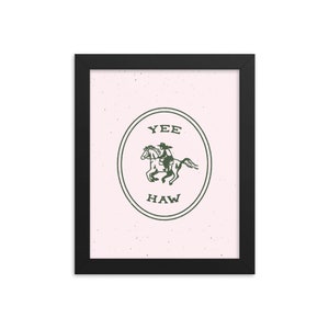 May include: A framed print with a pink background and a green illustration of a cowboy riding a horse inside a circle with the words "Yee Haw" written below the image.
