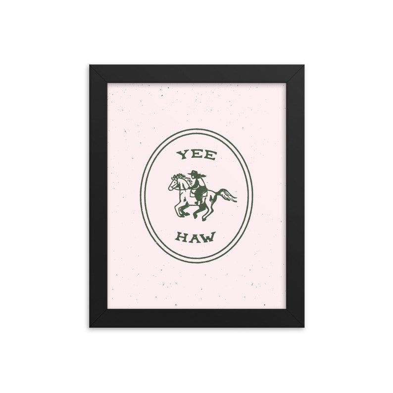 May include: A framed print with a pink background and a green illustration of a cowboy riding a horse inside a circle with the words "Yee Haw" written below the image.