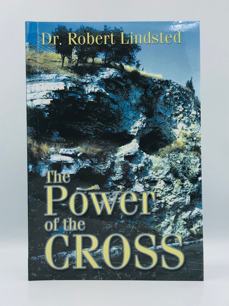 May include: Book cover with a blue and green rocky cliff with a cave opening. The title is "The Power of the Cross" in yellow text with the author "Dr. Robert Lindsted" in yellow text at the top.