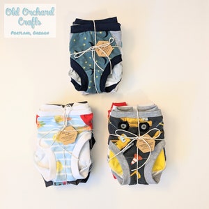 May include: Three stacks of colorful underwear for children. Each stack is tied with twine and a small brown tag that reads "Old Orchard Crafts, Handmade with Love, Portland, Oregon". The underwear features different patterns, including stars, moons, lemons, trucks, and construction cones.