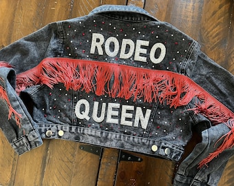 Könnte beinhalten: Eine schwarze Jeansjacke mit roten Fransen und Strasssteinen. Auf der Rückseite der Jacke steht in weißen Strasssteinen "Rodeo Queen" geschrieben.