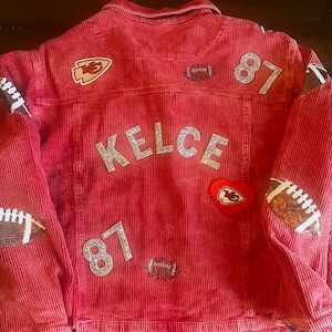 May include: Red corduroy jacket with a variety of patches, including a football, a heart with the Kansas City Chiefs logo, and the name "KELCE" and the number "87" in rhinestones.