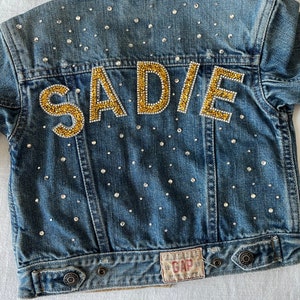 May include: A blue denim jacket with a gold glitter 'SADIE' design on the back. The jacket is covered in small, clear rhinestones.