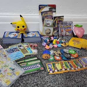 May include: A selection of Pokémon collectables, including trading cards, stickers, small figurines, and a Pikachu figure. The items are displayed on a surface, with card packs and a strawberry plush toy.