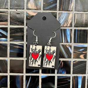 May include: A pair of silver earrings featuring a tarot card design. The card is the Five of Swords, depicting a red heart pierced by five swords. The card is set against a white background with black outlines.