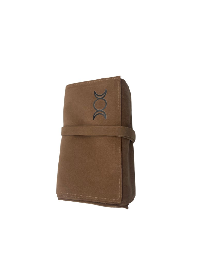 Puede incluir: Una bolsa de gamuza marr&oacute;n con un s&iacute;mbolo de triple luna en la parte delantera. La bolsa est&aacute; doblada y atada con una correa marr&oacute;n.