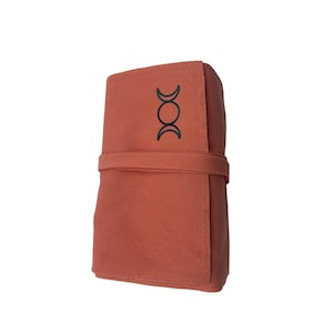 Puede incluir: Una bolsa de tela de color terracota con un s&iacute;mbolo de triple luna negro. La bolsa tiene una solapa que se pliega y se cierra con una correa.