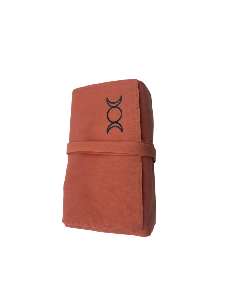 Puede incluir: Una bolsa de tela de color terracota con un s&iacute;mbolo de triple luna negro. La bolsa tiene una solapa que se pliega y se cierra con una correa.